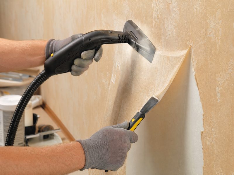 Professional hands carefully removing old wallpaper with a steamer and scraper